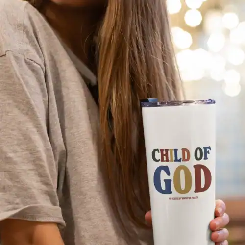 Child of God merch cup featuring colorful text and a religious message, held by a young woman with long brown hair, casual grey shirt, in a cozy indoor setting with warm bokeh lighting.